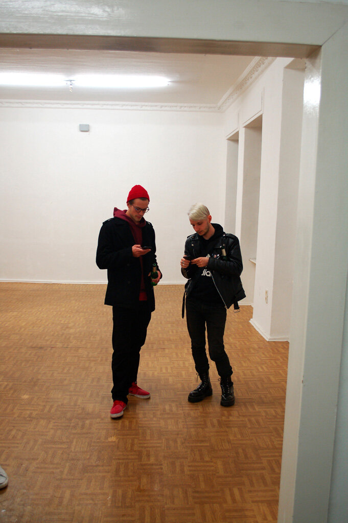 local.#non.access opening — two visitors on their phones in the middle of the empty exhibition space